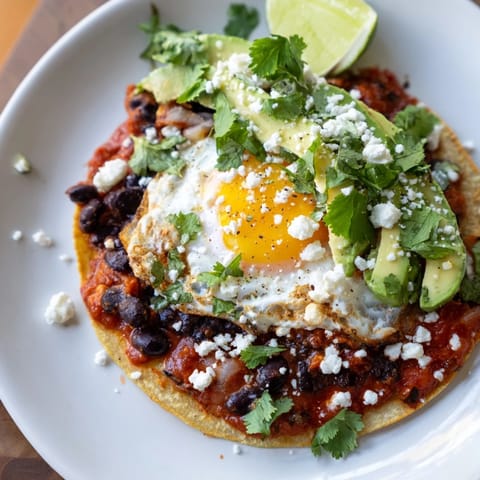 Warm corn tortillas layered with seasoned black beans, a fried egg, and a rich, simmered tomato sauce, finished with creamy avocado slices and tangy feta cheese for a hearty breakfast.