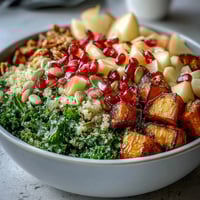 Brightly colored Kale Harvest Grain Bowl topped with roasted sweet potatoes, crisp apple, and ruby pomegranate seeds on a white plate.